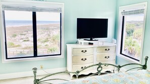 A bedroom with a bed, a dresser, a television, and two windows with a view of the beach.