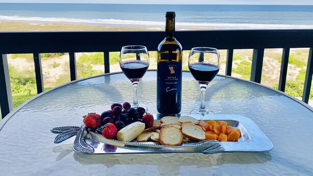 A table with a bottle of wine, two glasses of wine, and a plate of fruit and bread on a balcony overlooking the ocean.