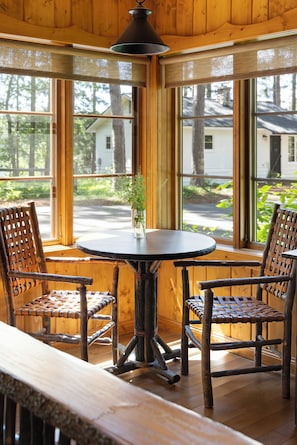 A wooden interior with a round table and two woven chairs, a hanging light fixture, and large windows with a view of a house and trees.