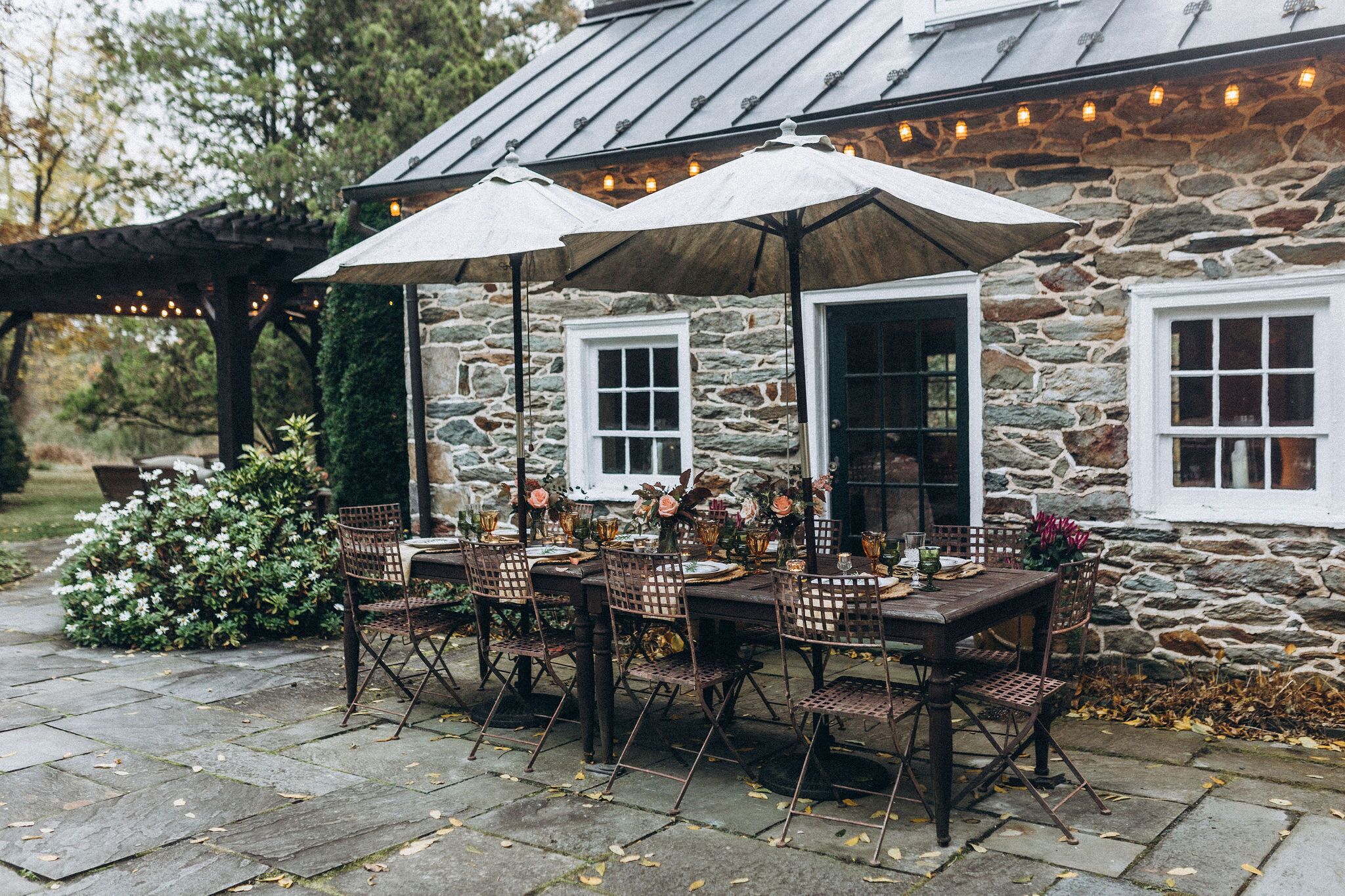 Een terras buiten met stenen muren, een houten tafel en stoelen, omgeven door groen en lichtjes.