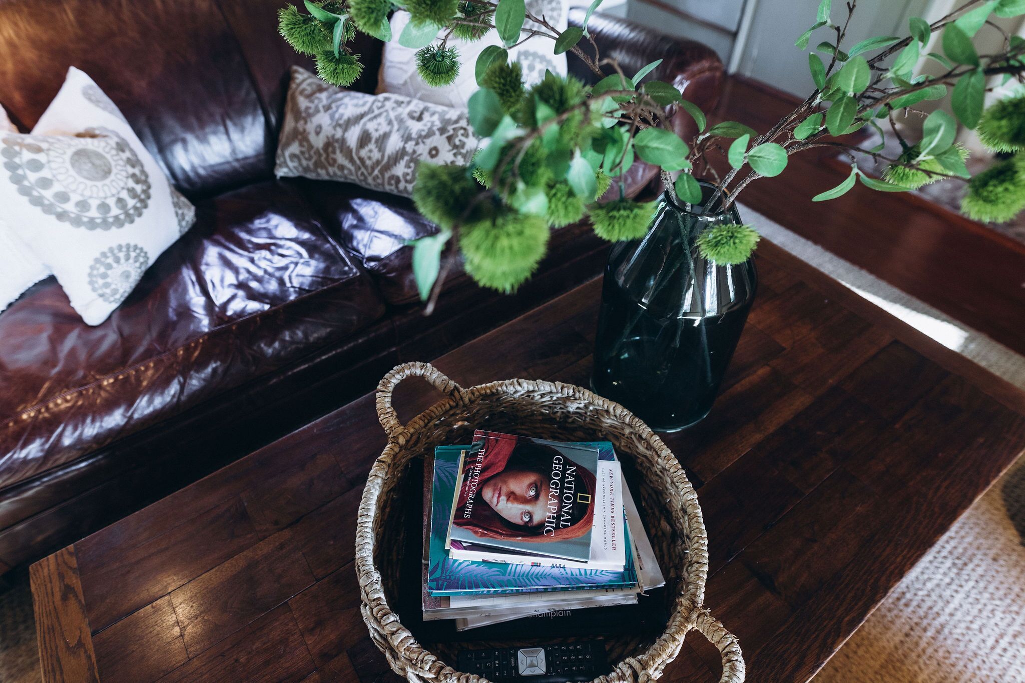 A wicker basket filled with books on a wooden table, a dark leather couch with patterned cushions, and a green plant in a glass vase.