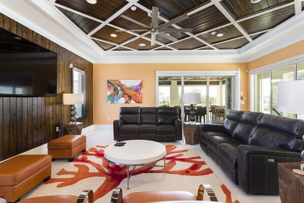 A spacious living area with a ceiling fan, a large flat-screen TV, a black leather sofa, a white coffee table, and a vibrant abstract painting on the wall.