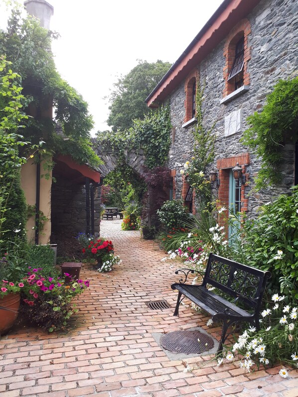 A brick-paved courtyard with a bench, surrounded by stone buildings and lush greenery.