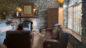 A cozy living room with a stone wall, a fireplace, a wooden cabinet, and a large window with blinds.