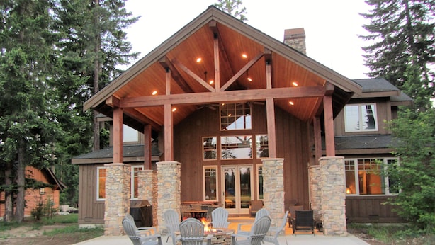 A large wooden house with stone pillars, a covered patio with outdoor furniture, and a fire pit.
