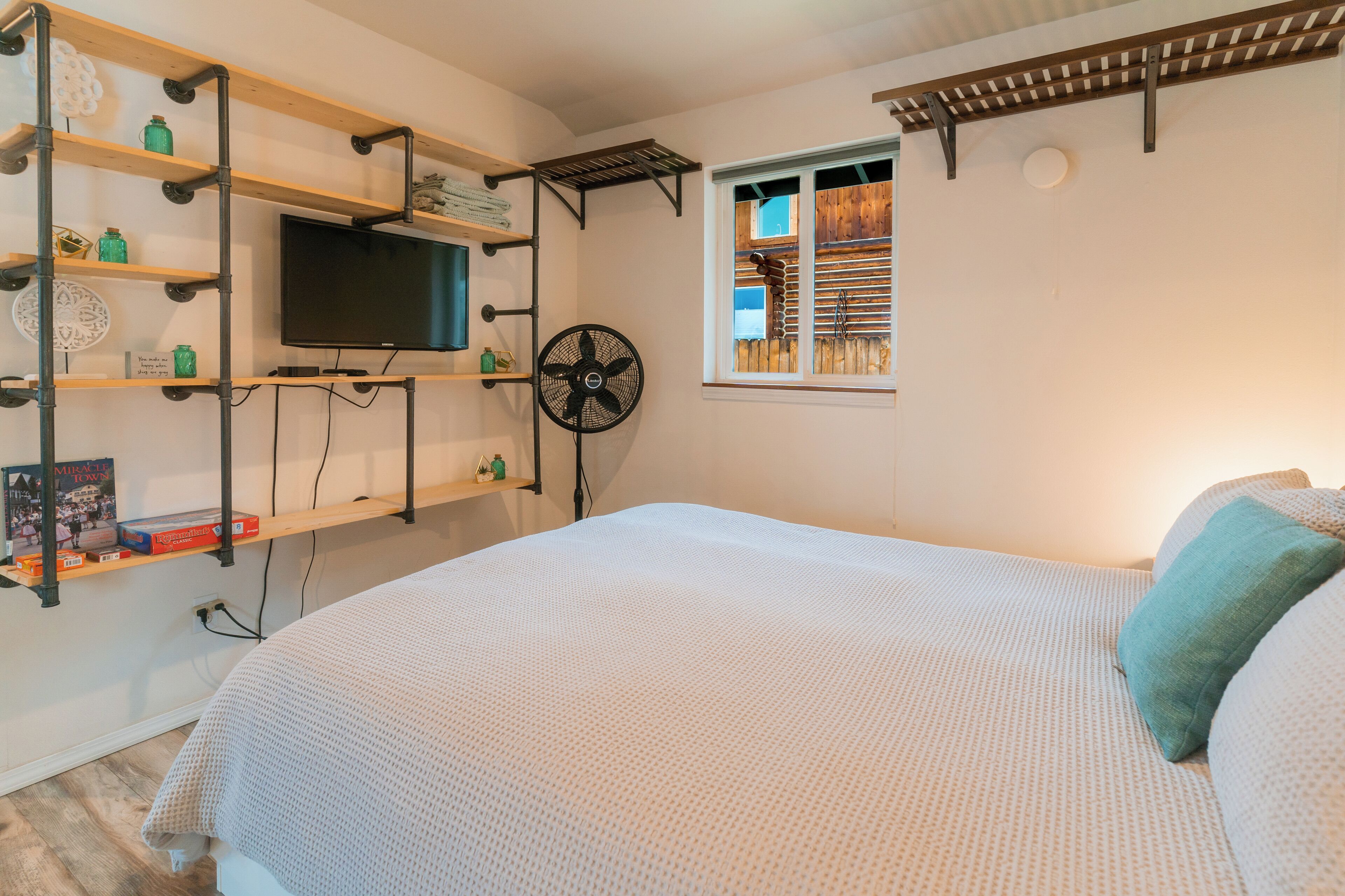 A bedroom with a bed, a TV mounted on a shelf, a window, and a fan.