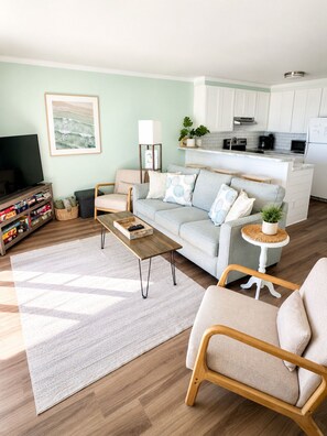 Una sala de estar moderna con sofá, mesa de centro y sillones, un área de cocina con armarios blancos y un televisor.