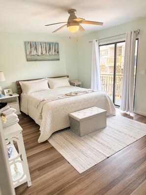Une chambre à coucher comprenant un lit, des tables de chevet, une commode, un ventilateur de plafond, une fenêtre avec des rideaux et une vue sur les bâtiments environnants.