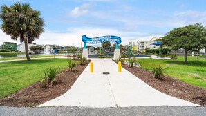 A pathway leading to a sign for Brandy Myers Memorial Playground with a water feature and greenery. - Oceanfront 2BR/2BA Pet-Friendly _ POOL!_ Walk to Boardwalk_ Private Beach Access (Carolina Beach)