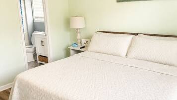 A bedroom with a large bed, a bench, a nightstand with a lamp, a painting on the wall, and a closet with a window.