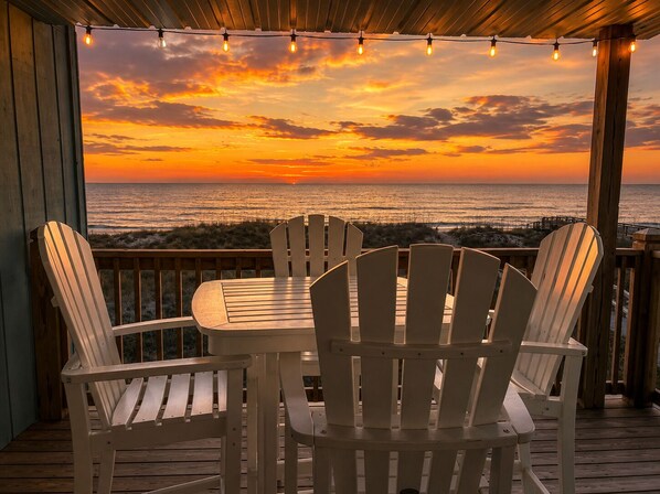 A wooden deck with white chairs and a table, overlooking a sunset over the ocean.