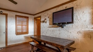 A room with a wooden table and benches, a television mounted on the wall, a window with blinds, and a door.