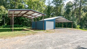 A gravel parking area with two metal sheds, one with a roof and one without, surrounded by trees.