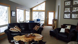 A living room with a black leather sofa, a wooden coffee table, and a cowhide rug. There are large windows with a view of trees. - Beautiful 4 Bedroom Renovated Home with A/C; 10 SHARC Passes (Sunriver)