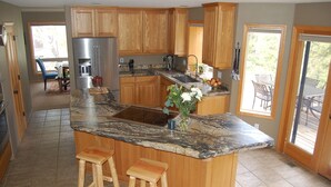 A kitchen with wooden cabinets, granite countertops, a stainless steel refrigerator, and a large island with stools. - Beautiful 4 Bedroom Renovated Home with A/C; 10 SHARC Passes (Sunriver)
