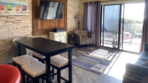 A cozy living area with a wooden table and chairs, a television mounted on a brick wall, and a sliding glass door leading to an outdoor area. - 405 Captain John Fripp Villa - Ocean Front - Ground Level (Fripp Island)