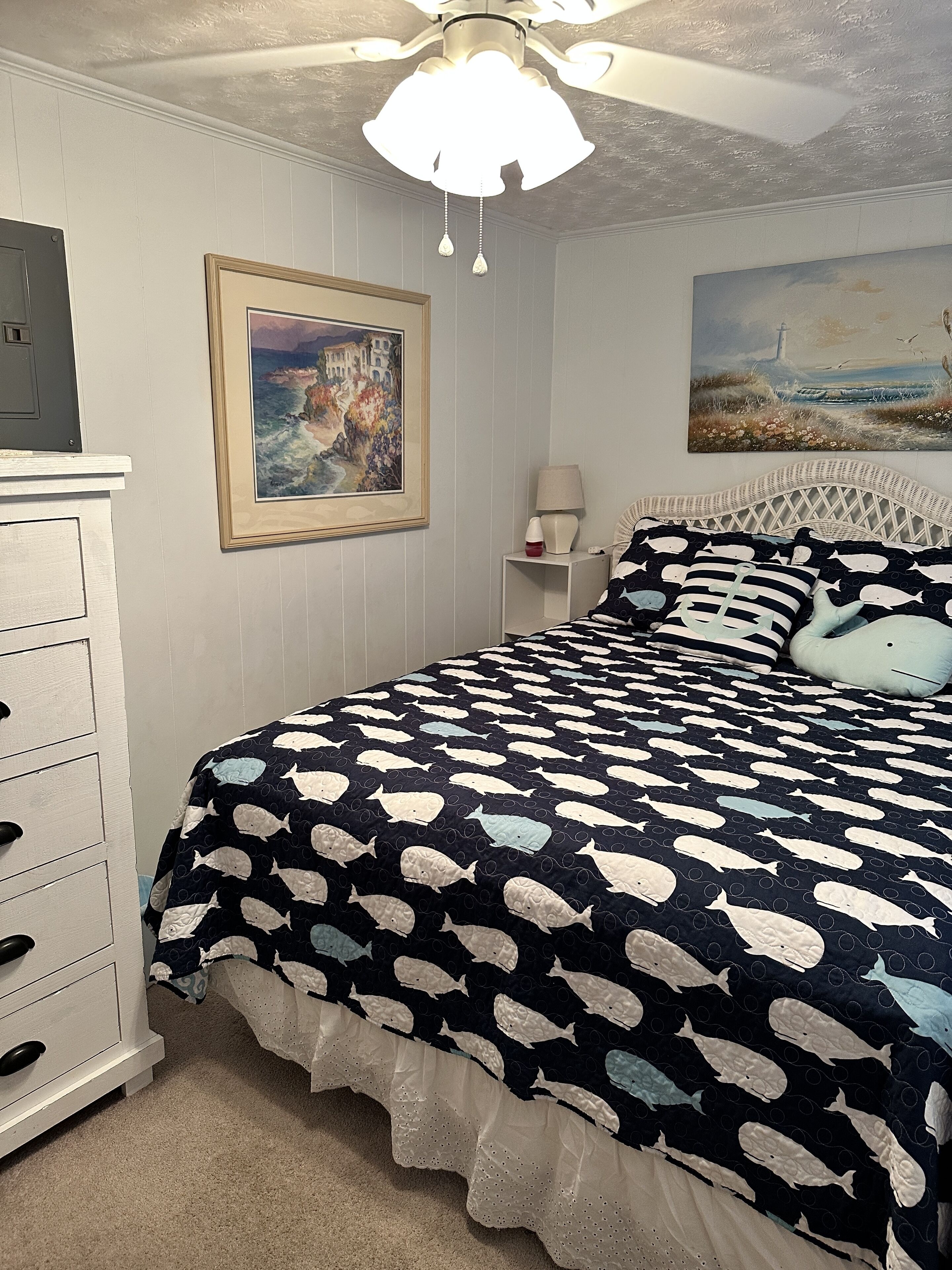 A bedroom with a bed, dresser, paintings, and a ceiling fan.