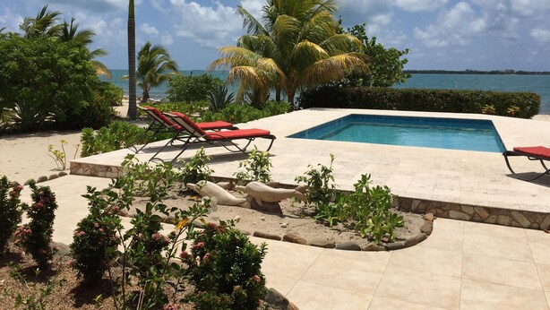 Zona de piscina con dos sillones reclinables rojos, rodeada de plantas tropicales y palmeras, con vista al océano.