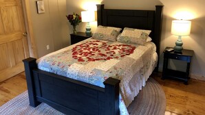 A neatly made bed with a floral bedspread and a heart-shaped arrangement of rose petals. There are two bedside tables with lamps, a framed picture on the wall, and a wooden door. - Spacious Bobcat Cabin, 30 mins from Hocking Hills (Nelsonville)