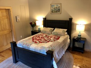 A neatly made bed with a floral bedspread and a heart-shaped arrangement of rose petals. There are two bedside tables with lamps, a framed picture on the wall, and a wooden door.
