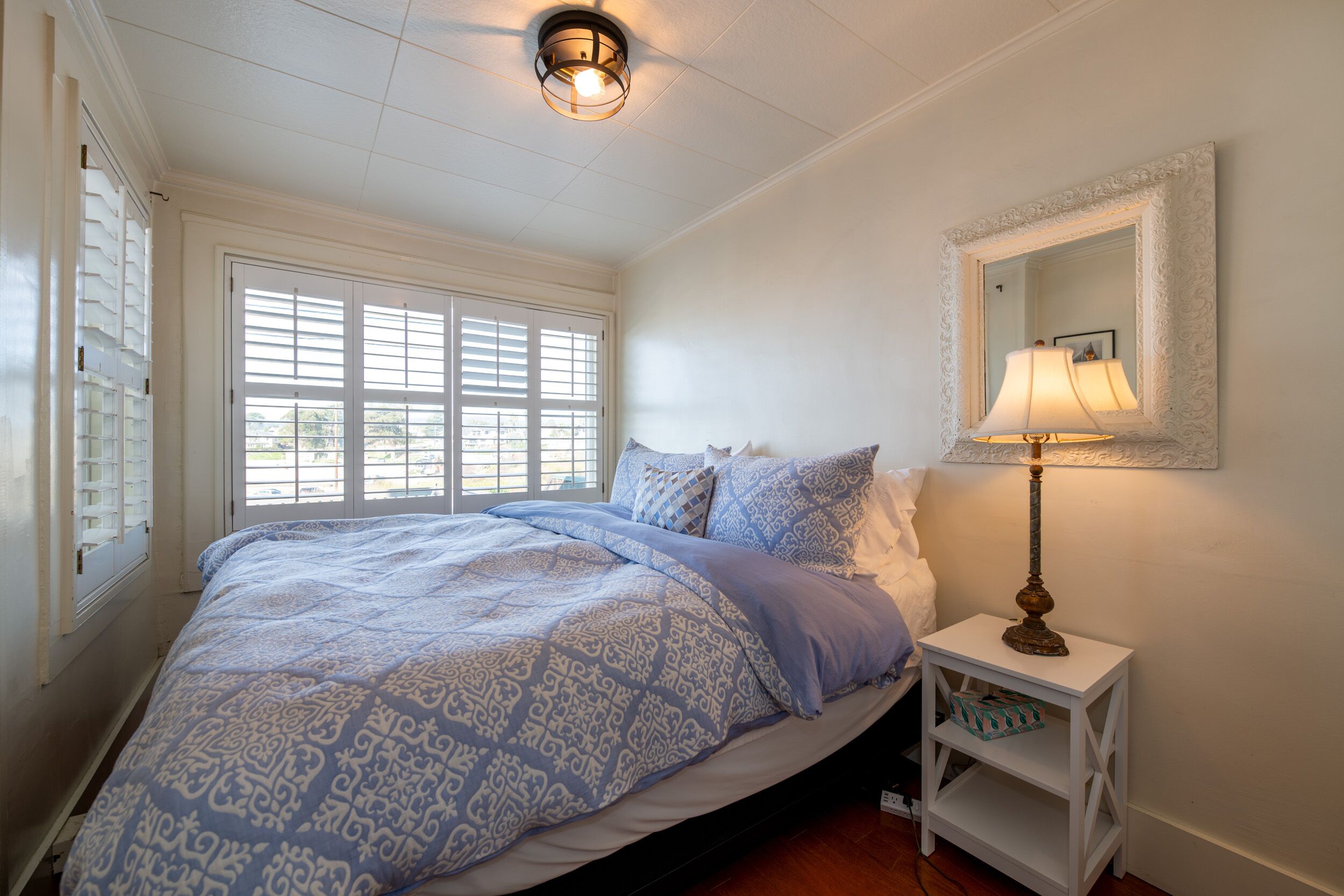 A bedroom with a bed, a nightstand with a lamp, a window with blinds, and a mirror on the wall.