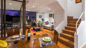 A modern living room with a dining area, a staircase, and a view of the city at dusk. - Riverfront condo - Amazing views in Cuenca's Historic District  (Cuenca)