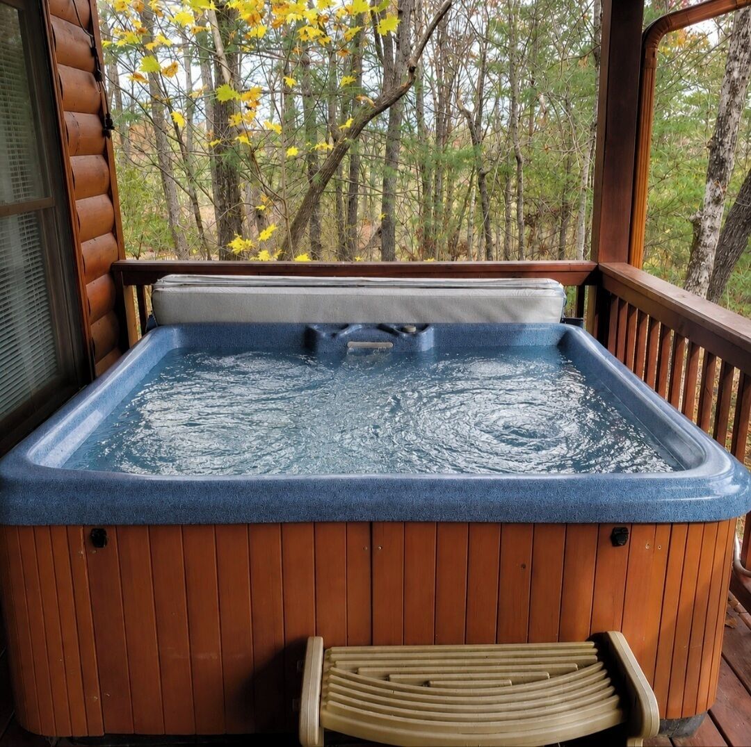 A hot tub with a wooden deck and a bench, surrounded by trees.