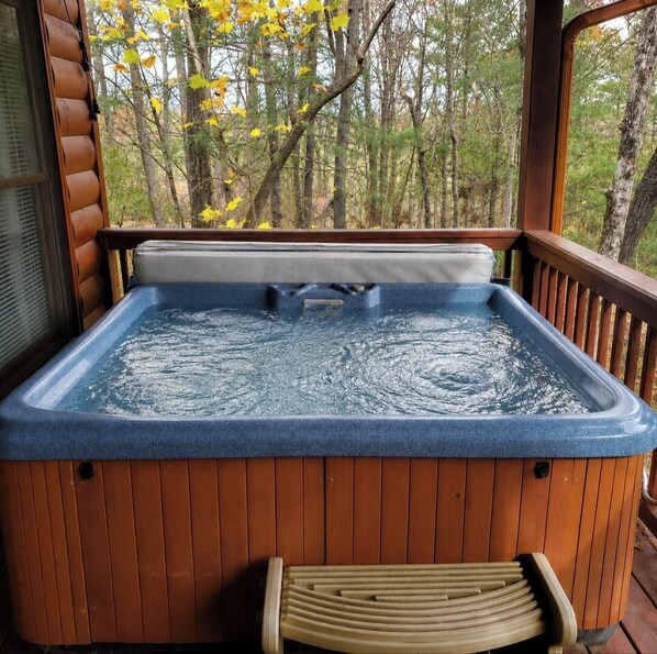 A hot tub with a wooden deck and a bench, surrounded by trees.