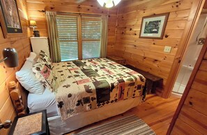A wooden cabin room with a bed, a nightstand, a lamp, a window with blinds, and a framed picture on the wall. - WHAT A VIEW from the HOT TUB! Ping-pong & Pac-Man game room! (Blue Ridge)
