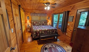 A wooden cabin bedroom with a bed, bedside lamps, a ceiling fan, and a window with curtains.