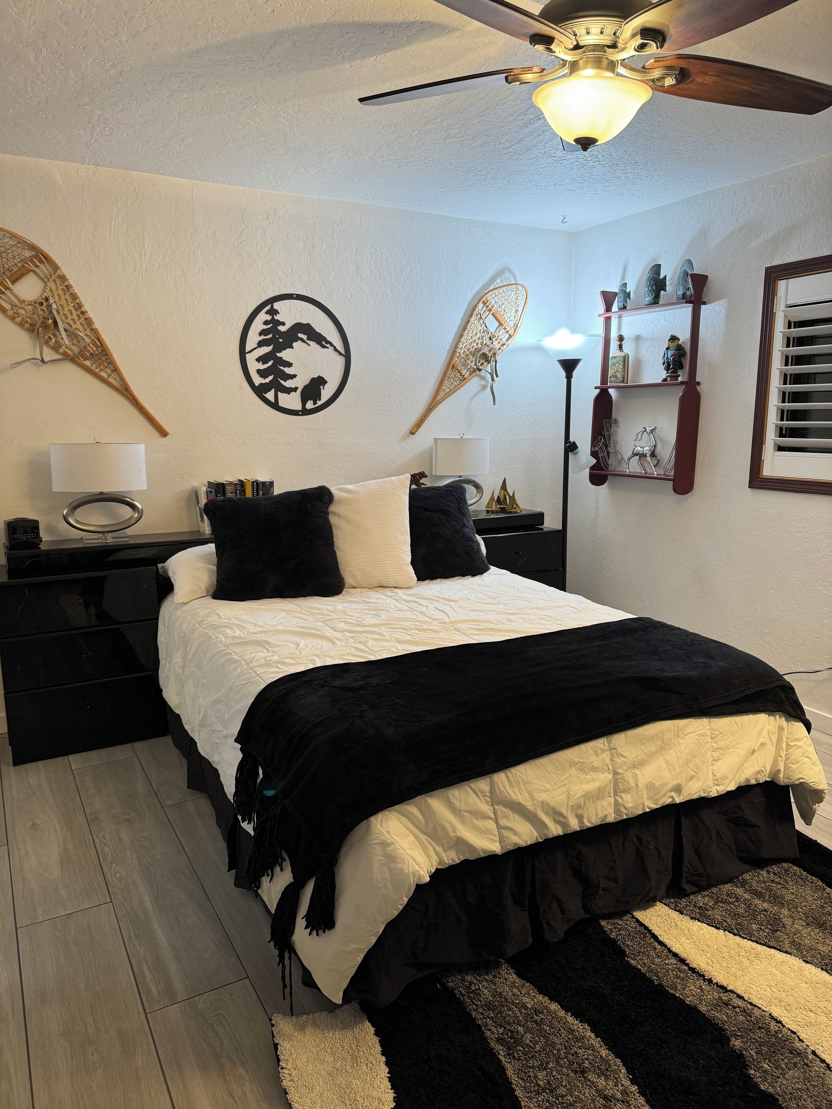 A bedroom with a bed, a ceiling fan, a nightstand, a lamp, wall art, and a shelf with decorative items.