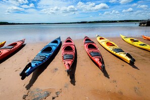 A row of kayaks lined up on a sandy beach with a calm body of water and a dock in the background. - Oceanfront Getaway | Walk to Harbour | Quiet & Scenic (North Rustico)