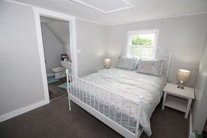 A bedroom with a white metal bed frame, floral bedding, two bedside tables with lamps, a window, and a view of the bathroom. - Oceanfront Getaway | Walk to Harbour | Quiet & Scenic (North Rustico)