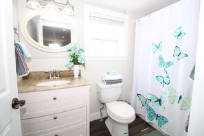 A bathroom with a white sink, a toilet, a mirror, and a shower curtain with butterfly designs. - Oceanfront Getaway | Walk to Harbour | Quiet & Scenic (North Rustico)