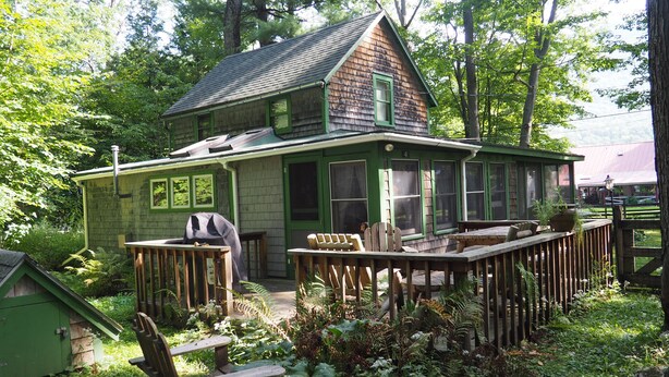A green house with a deck, surrounded by lush greenery. - Classic Vermont Cabin on 2.5 acres Lake Frontage (Salisbury)