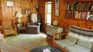 A cozy room with a wooden interior, a round coffee table with a book, a sofa, a chair, a small table with a lamp, and a bookshelf filled with books. - Classic Vermont Cabin on 2.5 acres Lake Frontage (Salisbury)