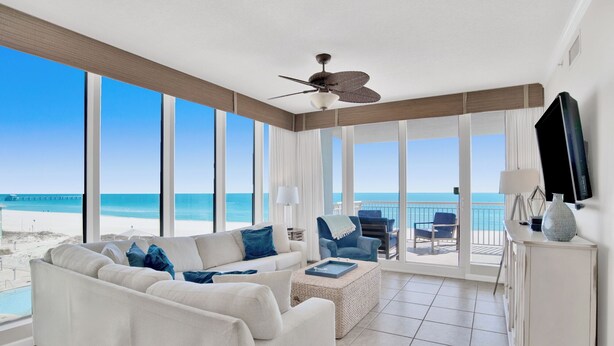 A spacious living room with a large window overlooking the beach, a ceiling fan, and a TV mounted on the wall.