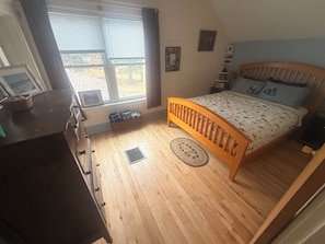 A bedroom with a wooden bed, a dresser, a window with blinds, and a view of the outdoors.