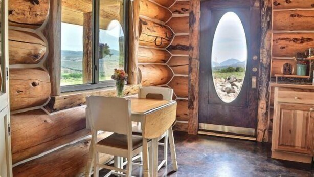 Interior de una cabaña de troncos con comedor, un ventanal y una puerta que ofrece vistas del exterior.