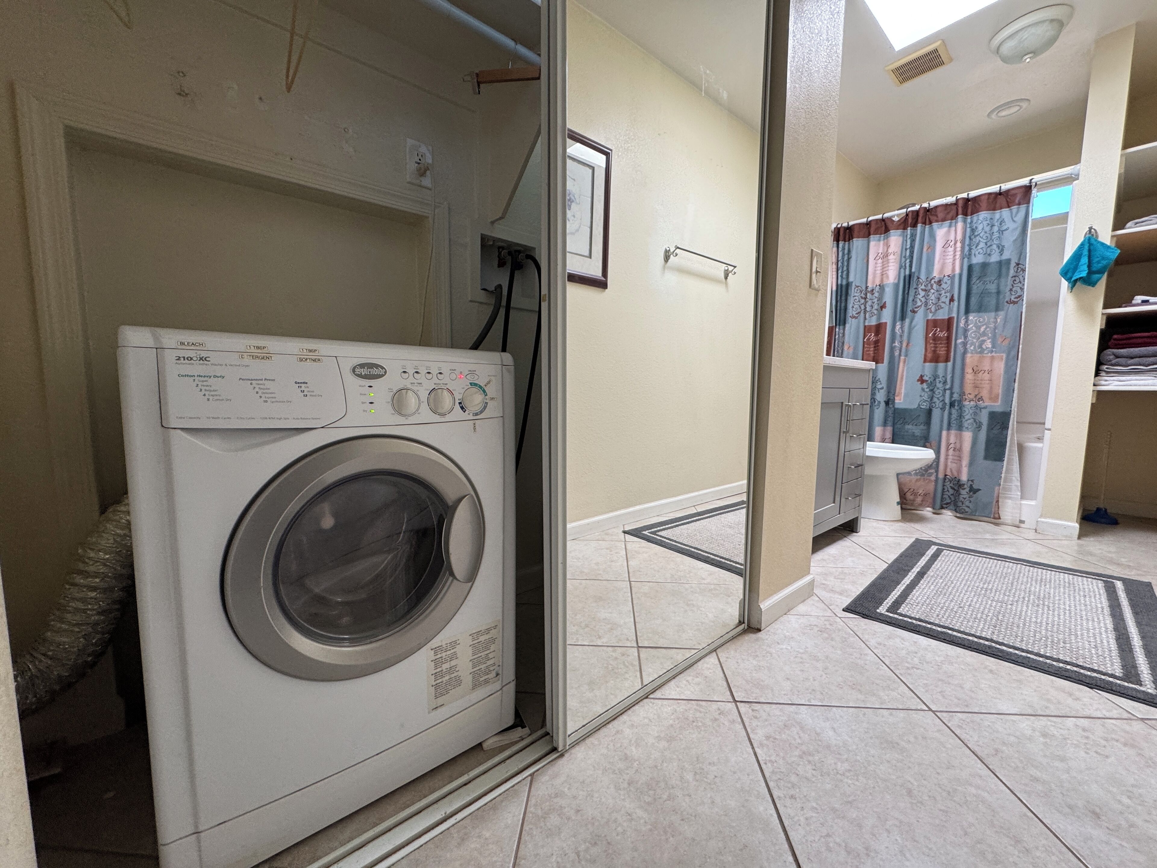 Une machine à laver à chargement frontal se trouve dans une buanderie dont le sol est carrelé, et on peut apercevoir une salle de bain à travers une porte ouverte.