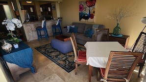 Una sala de estar con sofá, reposapiés y una mesa de comedor con sillas. Se observa una cocina al fondo.
