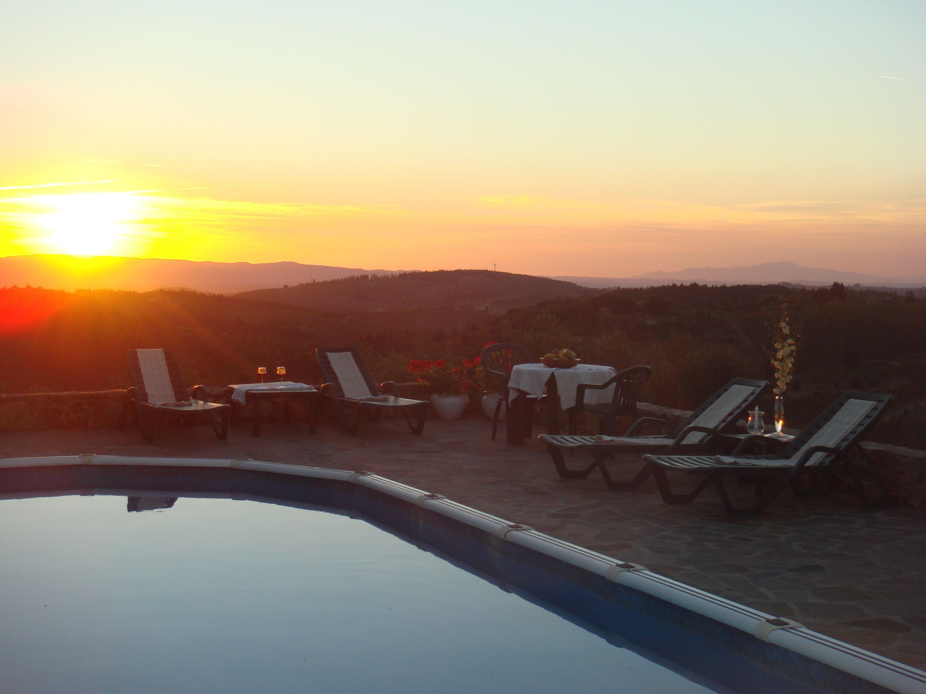 Zona junto a la piscina con sillones de descanso y una mesa para dos, con vista a un hermoso atardecer.