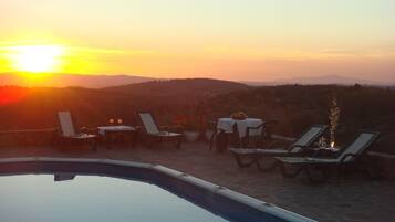 Zona junto a la piscina con sillones de descanso y una mesa para dos, con vista a un hermoso atardecer.