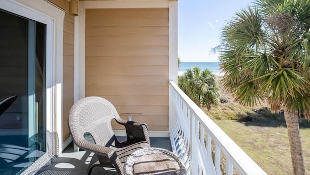 A balcony with wicker chairs, a glass door, and a view of the beach and palm trees.