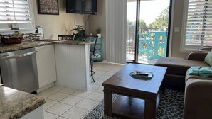 A compact kitchen with a built-in dishwasher, a flat-screen TV mounted above the counter, and a dining area with a table and chairs. - Admiral’s Row B2 - 2nd floor corner condo.  Clean, comfy and relaxing (Hilton Head Island)