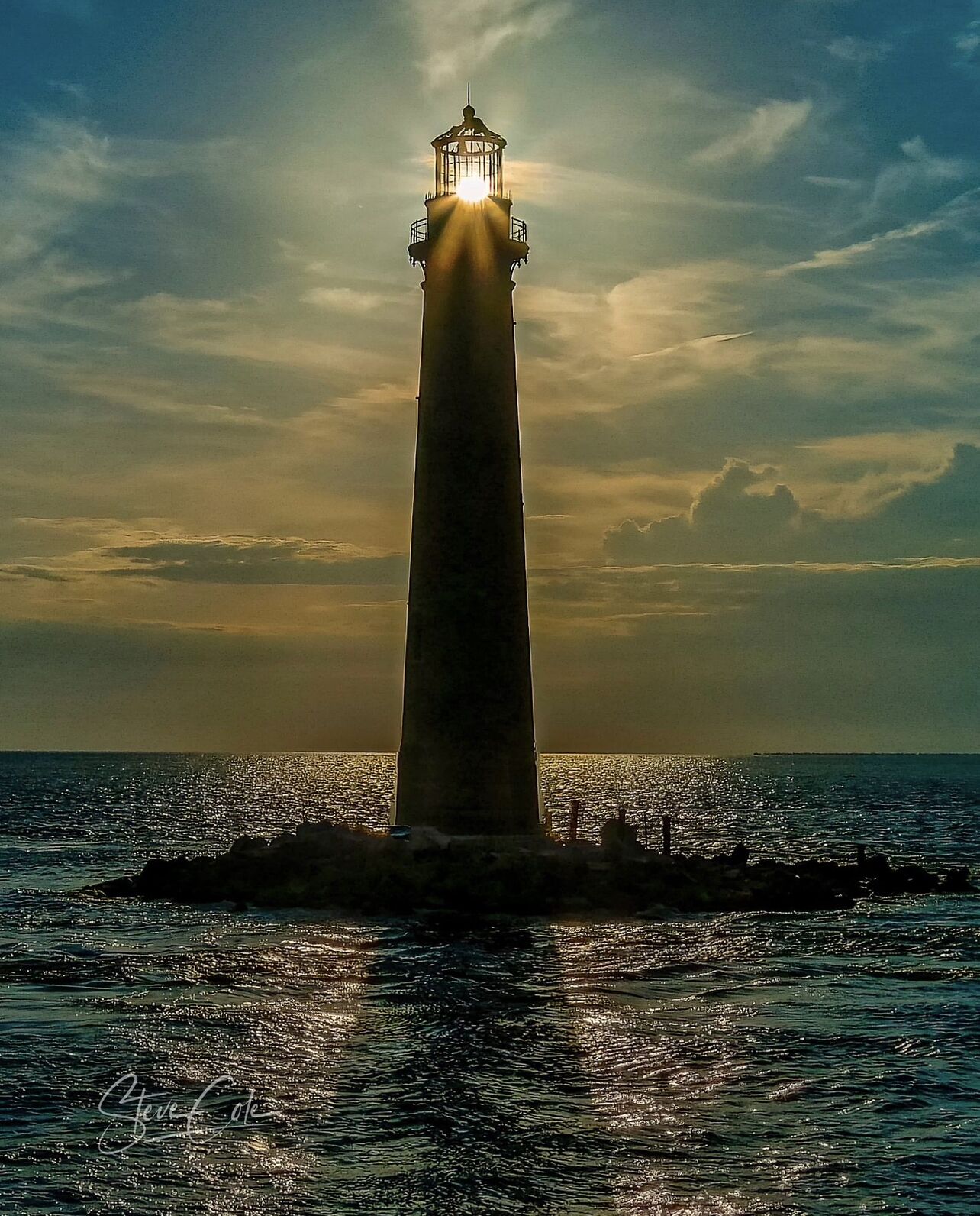 A lighthouse with a glass lantern room, surrounded by water and a cloudy sky.