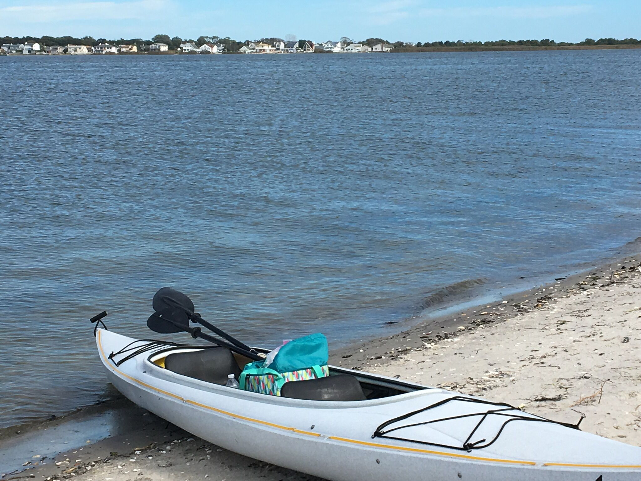 A kayak with a paddle and a bag on a sandy shore by a calm body of water.