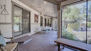 A covered patio area with a dining table and chairs, a bench, and a large window offering a view of the outdoors.