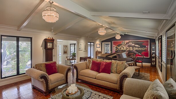 A spacious living room with a grand piano, elegant chandeliers, and a large window offering a view of the outdoors.