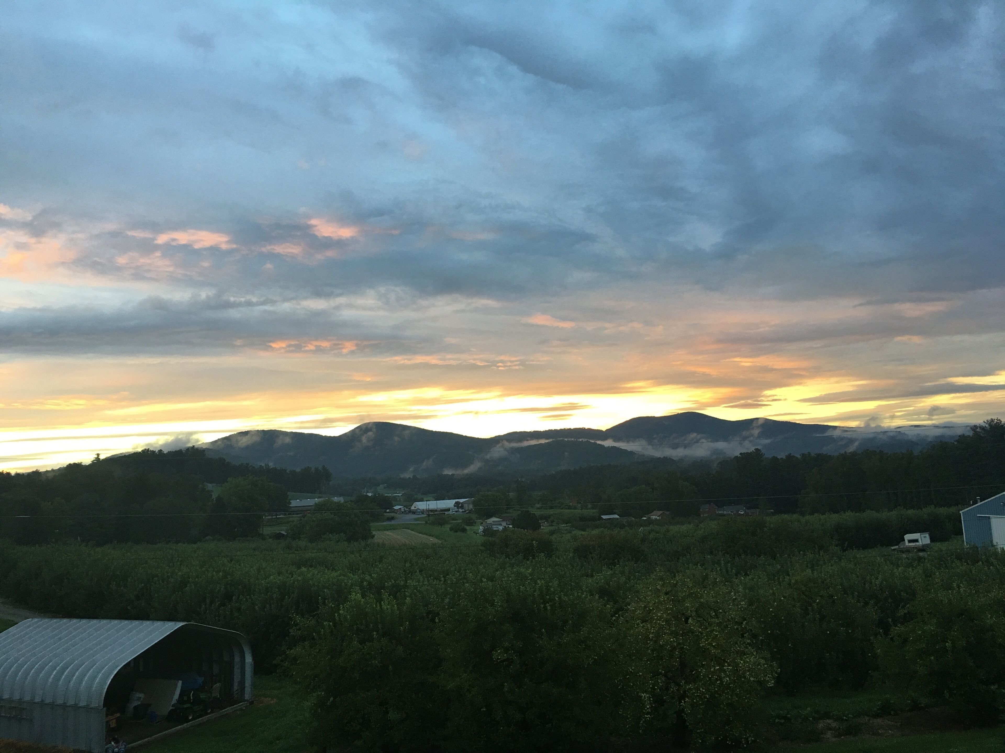 Paesaggio rurale con un tramonto, montagne e un piccolo edificio in primo piano.
